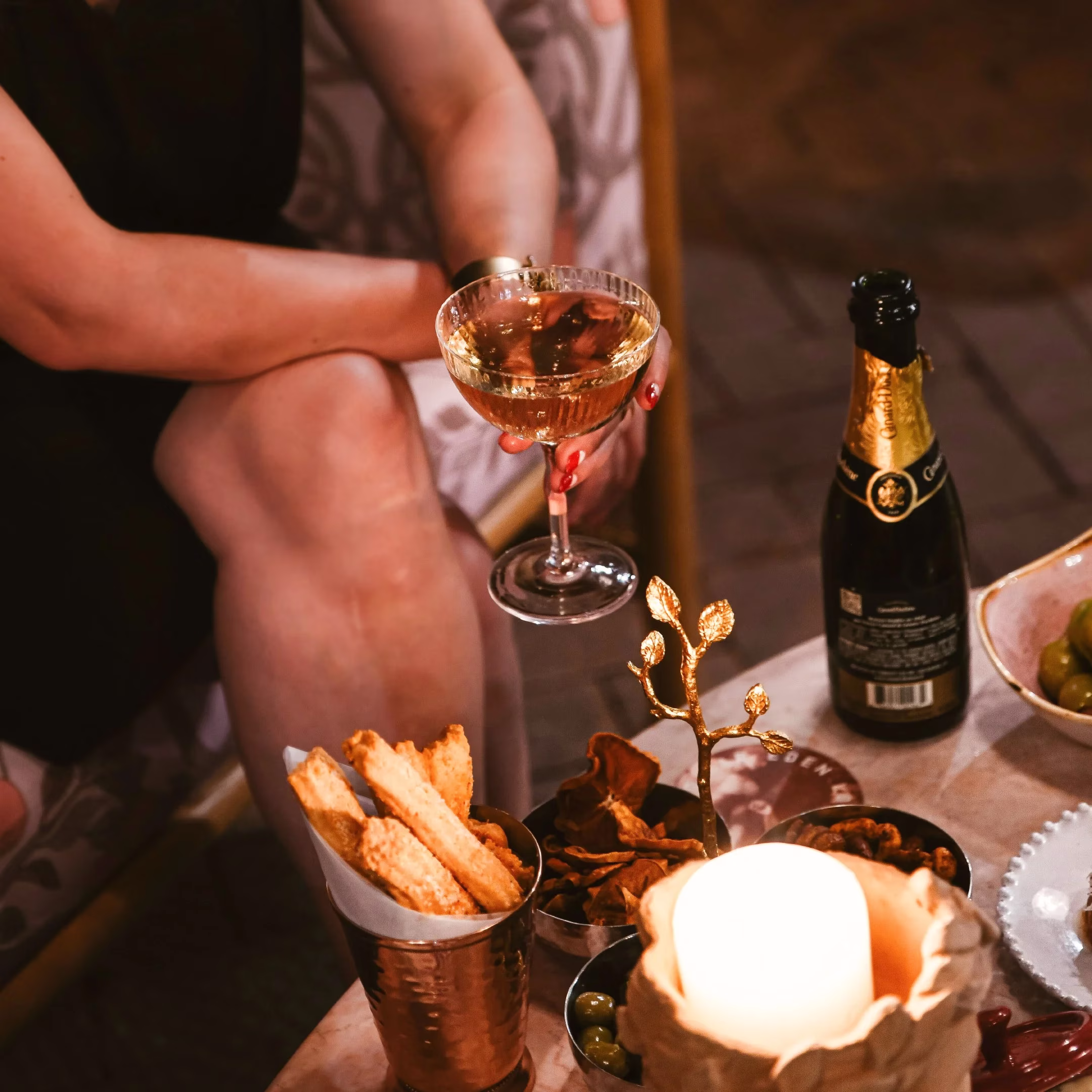 Person holding champagne glass, snacks, champagne bottle, lit candle.