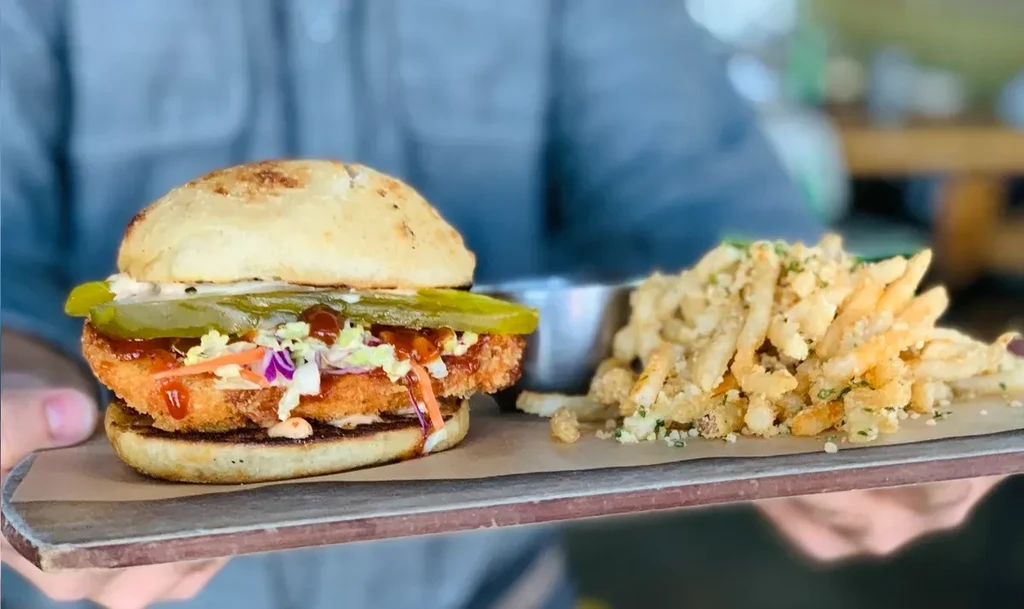 Hands hold a burger, fries, coleslaw, and pickle on a wooden board.
