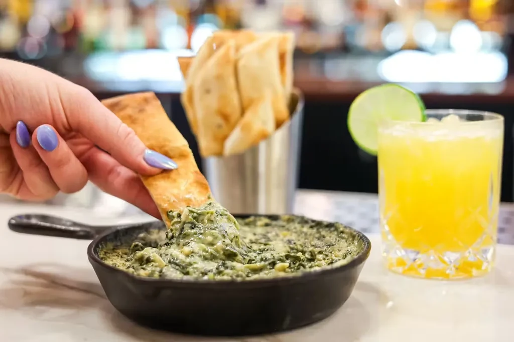 Hand dips pita into creamy green dip in cast iron skillet.