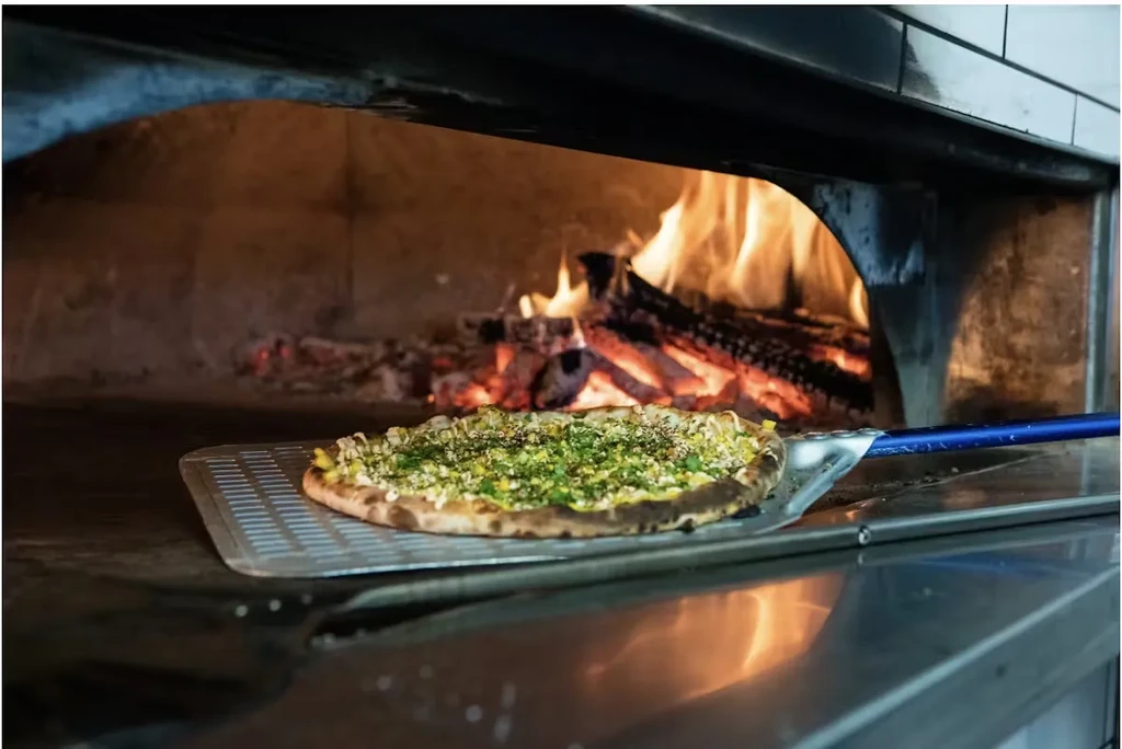 Fresh pizza on a peel near a roaring wood-fired oven.
