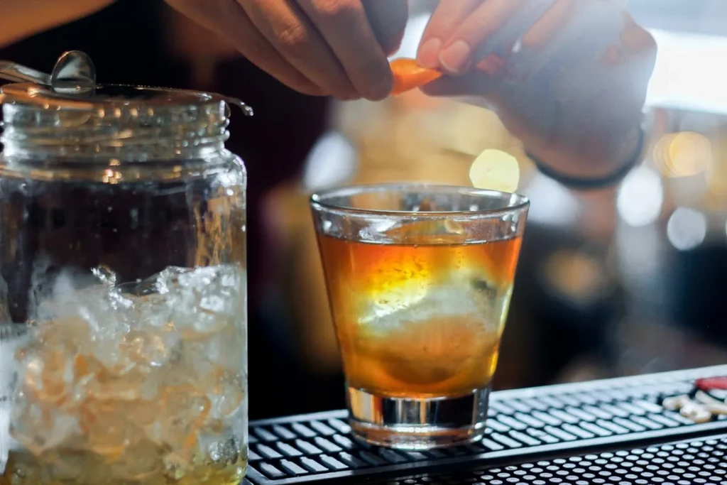 Close-up: Hands garnish amber cocktail with orange peel at bar.