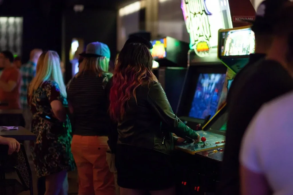 People play brightly lit arcade games in a dim, vibrant venue.