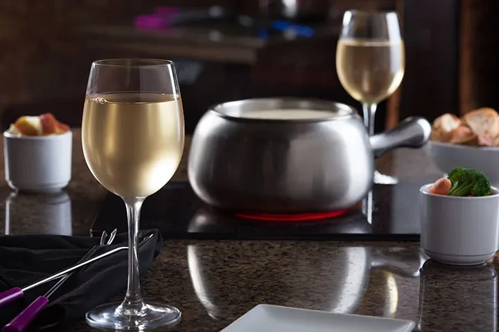 Close-up of fondue pot, wine, forks, and dippers on dark stone.