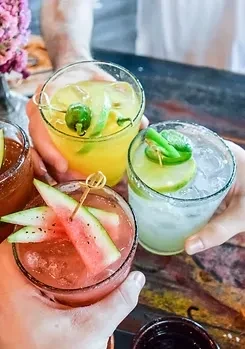 Three hands toast four colorful cocktails with unique garnishes.