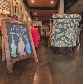 Rustic chalkboard "Let's Unwind" with bottles, beside a floral armchair.