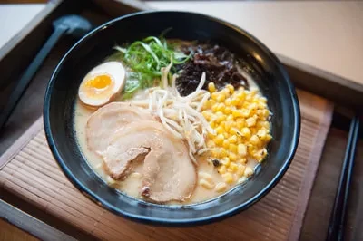 A dark ramen bowl with chashu, soft-boiled egg, corn, and greens.