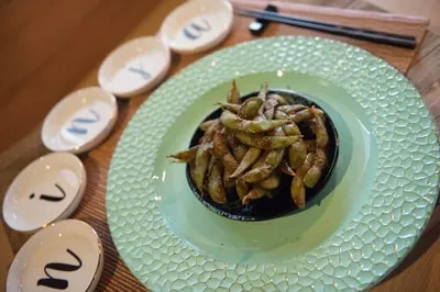 Close-up of spiced edamame, textured teal plate, and five letter dishes.