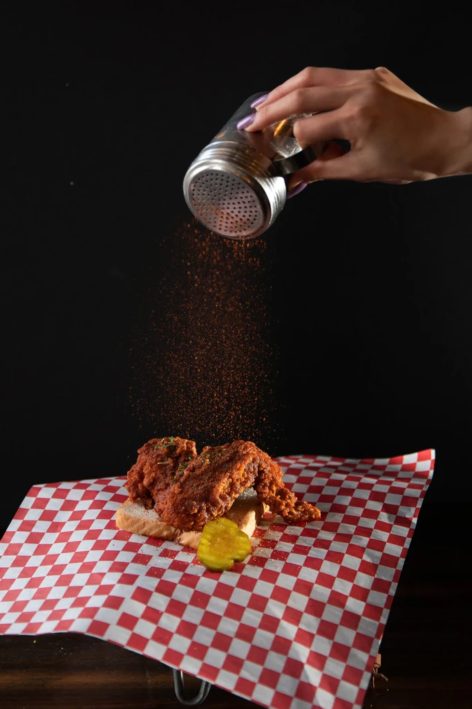 Close-up of fried chicken on bread with pickle, being seasoned.