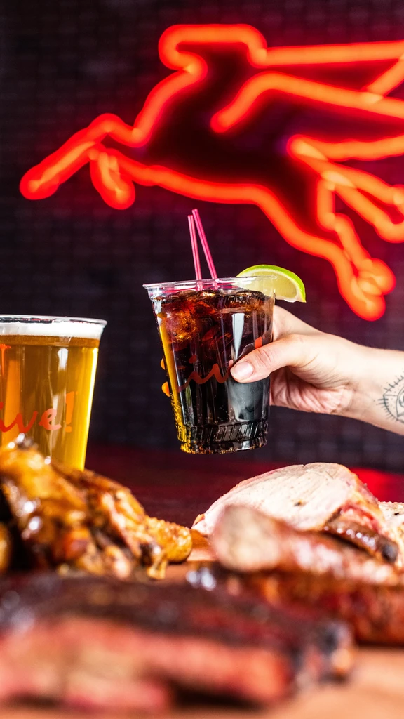 Hand holds soda with lime; beer, barbecue, and red neon sign in background.