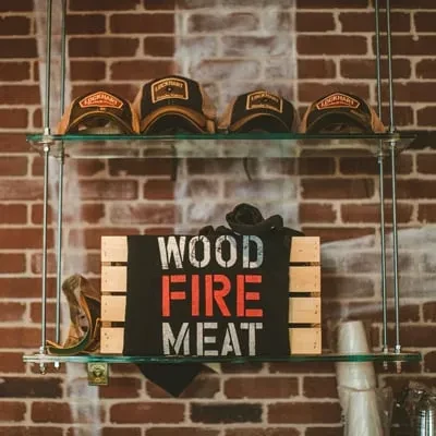 Brick wall shelves display caps, 'WOOD FIRE MEAT' sign, and cups.