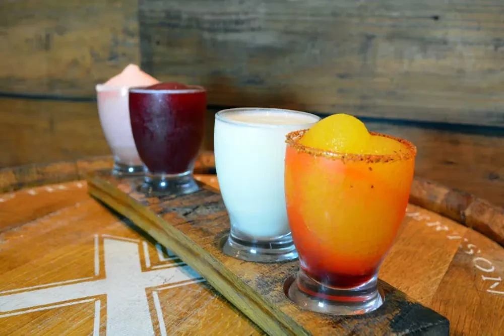 Four colorful frozen drinks, one with a spiced rim, on a wooden tray.