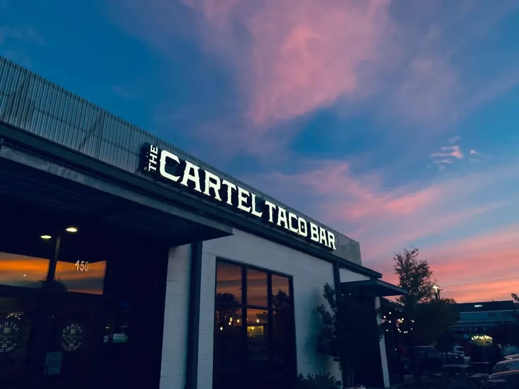Illuminated "THE CARTEL TACO BAR" exterior under a dramatic sunset sky.