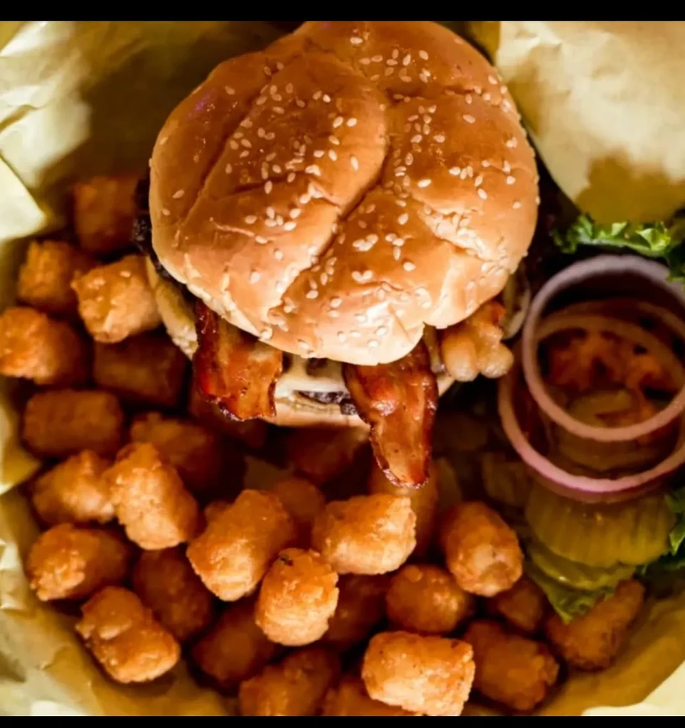 Large bacon cheeseburger and tater tots with garnishes in a basket.