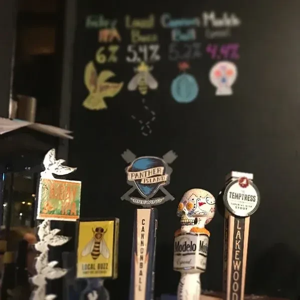 Close-up: varied beer tap handles on a bar counter, blurred chalkboard menu.