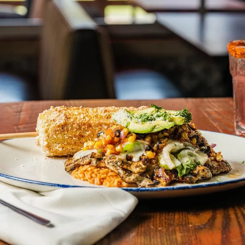 Plate of grilled meat, orange rice, salsa, avocado, and an elote.