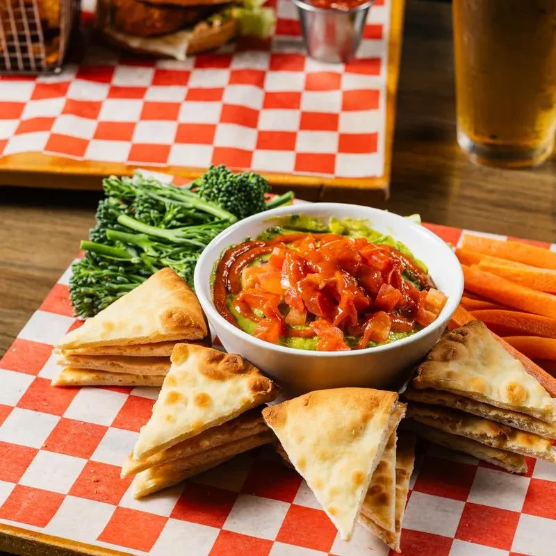 Guacamole bowl, pita bread, broccolini, and carrots on a checkered board.