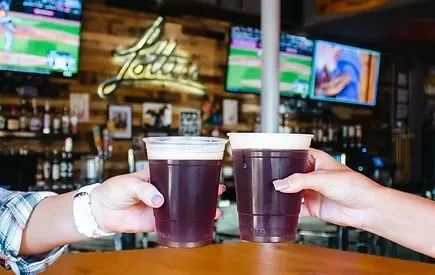 Two hands toast dark beverages in plastic cups at a sports bar.