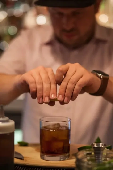 Bartender's hands squeezing citrus peel into a dark drink glass.