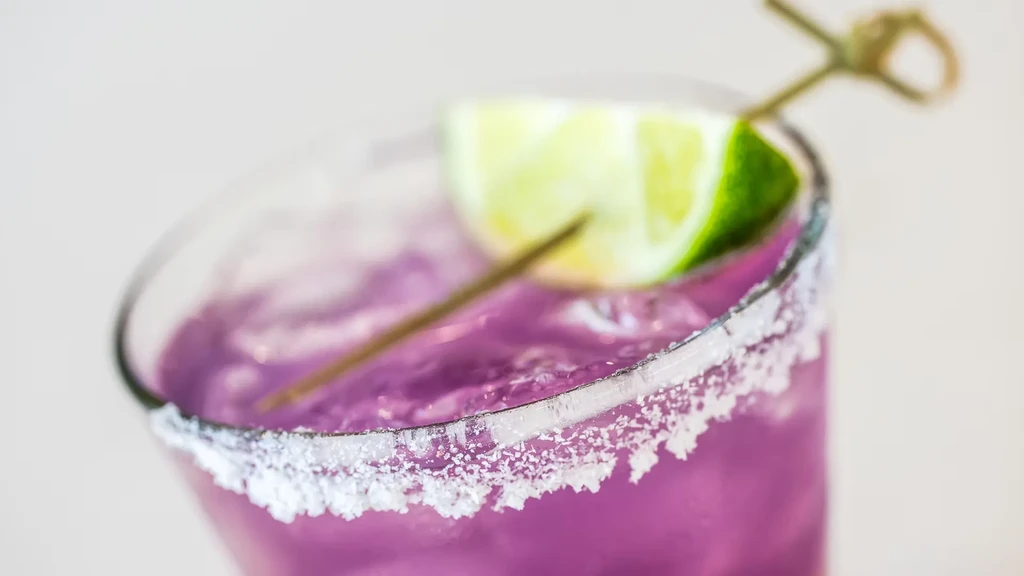 Close-up of a purple cocktail in a salt-rimmed glass with lime.