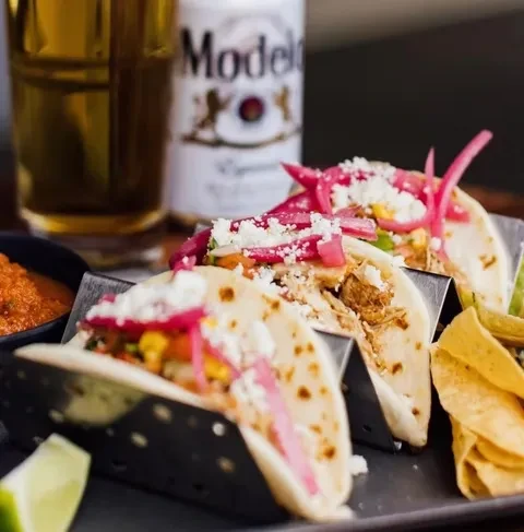 Three tacos with pickled onions, cheese, chips, salsa, and Modelo beer.