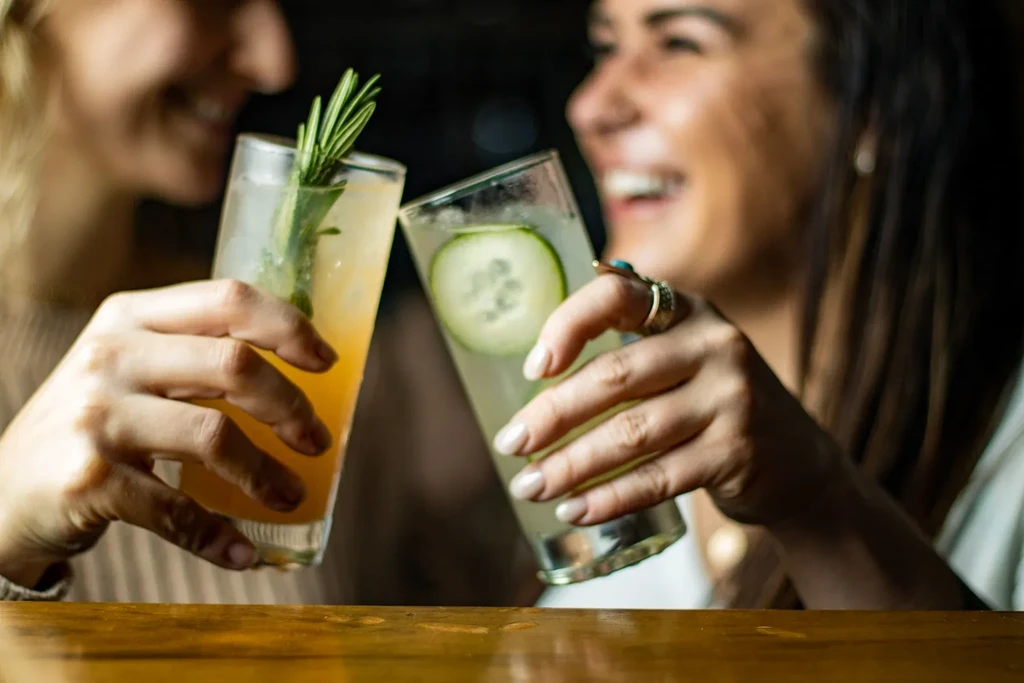 Two people toast amber and clear cocktails with rosemary and cucumber.