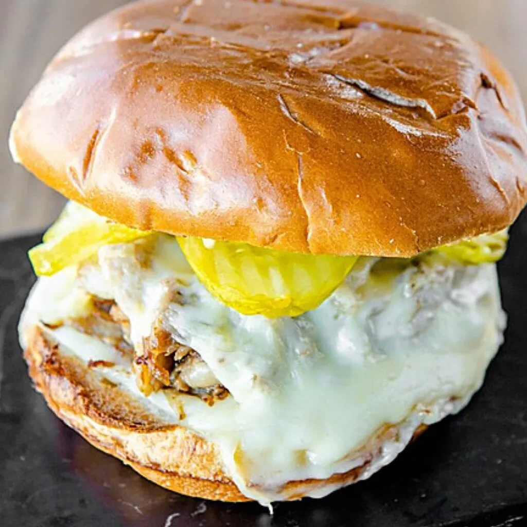 Close-up pulled meat sandwich with melted cheese and pickle slices.