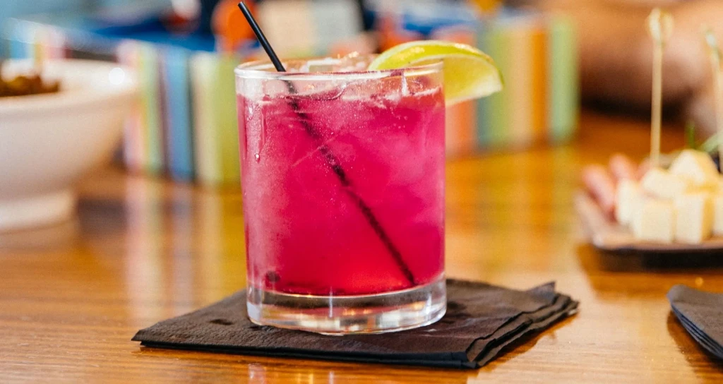 Magenta drink with lime wedge and black straw on a wooden table.