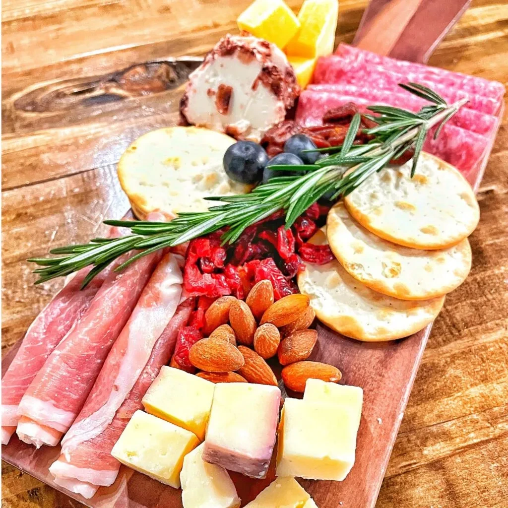Charcuterie board on wooden paddle with meats, cheeses, fruits, nuts, rosemary.
