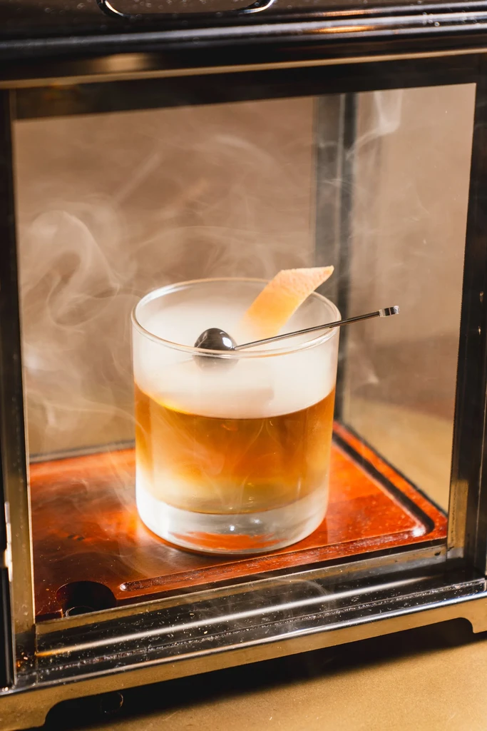Amber cocktail with garnish inside a smoking box with white smoke.
