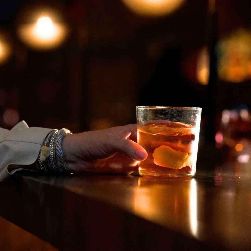 Hand with bracelets holds amber cocktail with ice and citrus.