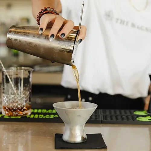 Hands pour golden-brown liquid from silver shaker into flared glass.