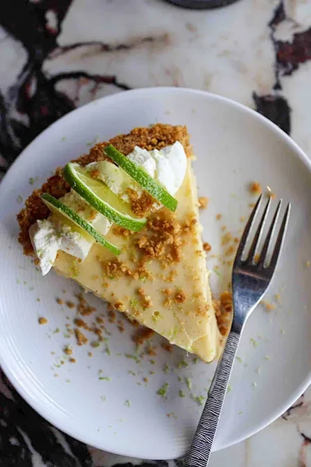 Key Lime Pie slice with whipped cream, lime, and fork on marbled counter.