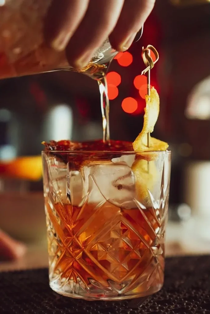 Hand pouring amber cocktail with ice, citrus, and dark garnishes.