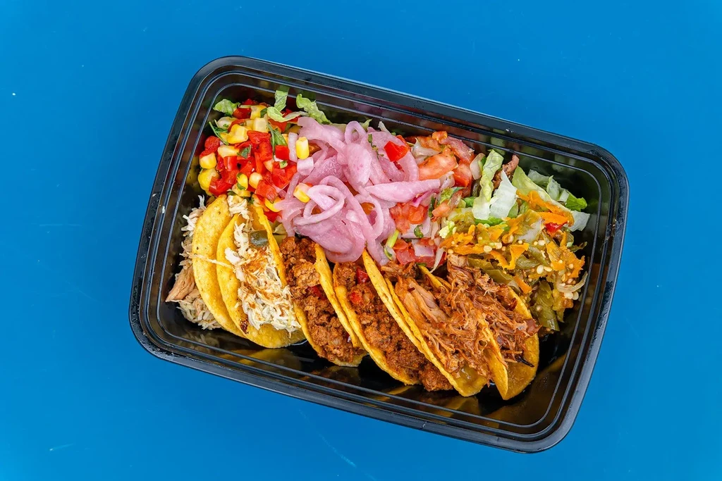 Overhead view of six tacos and toppings in a black takeout container.