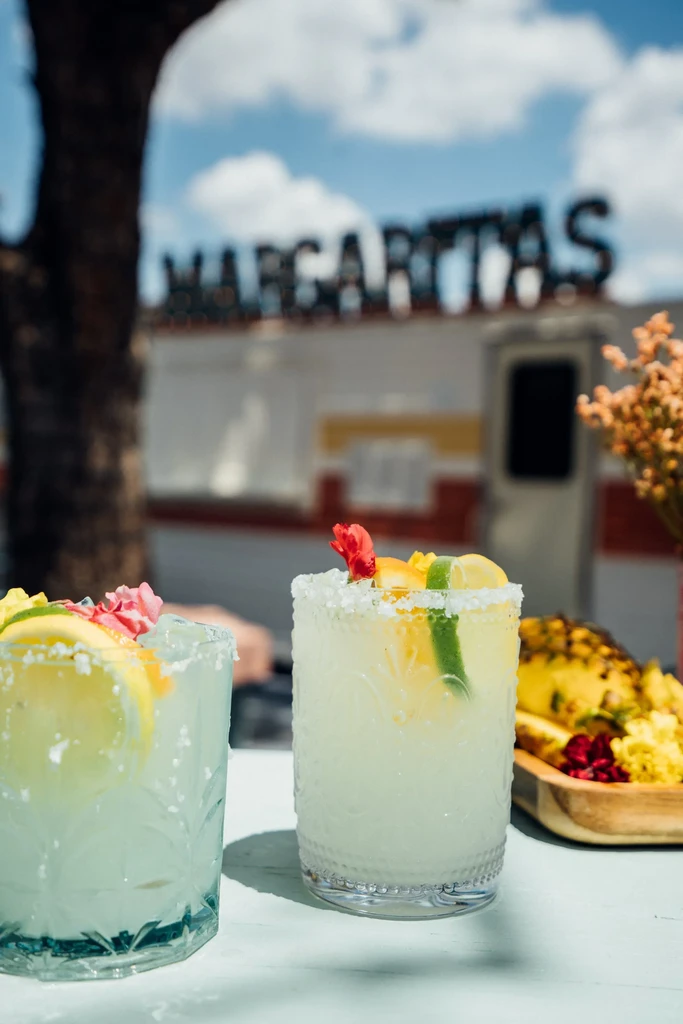 Two margaritas with flowers and citrus near a "MARGARITAS" sign.