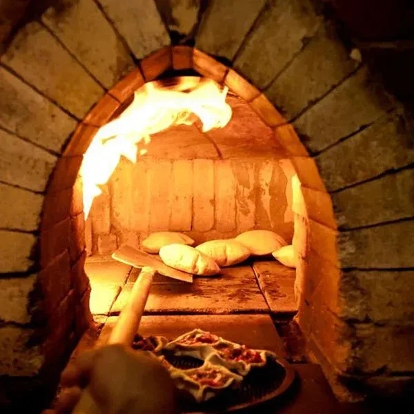 Hand holds peel with pastries baking in firelit brick oven.