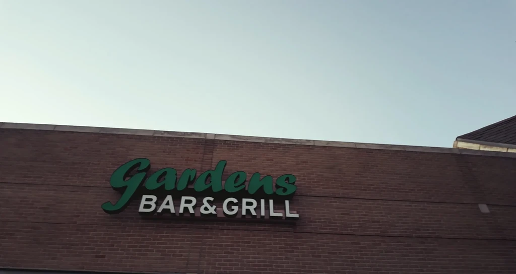 Red brick building with a 'Gardens BAR & GRILL' sign under blue sky.