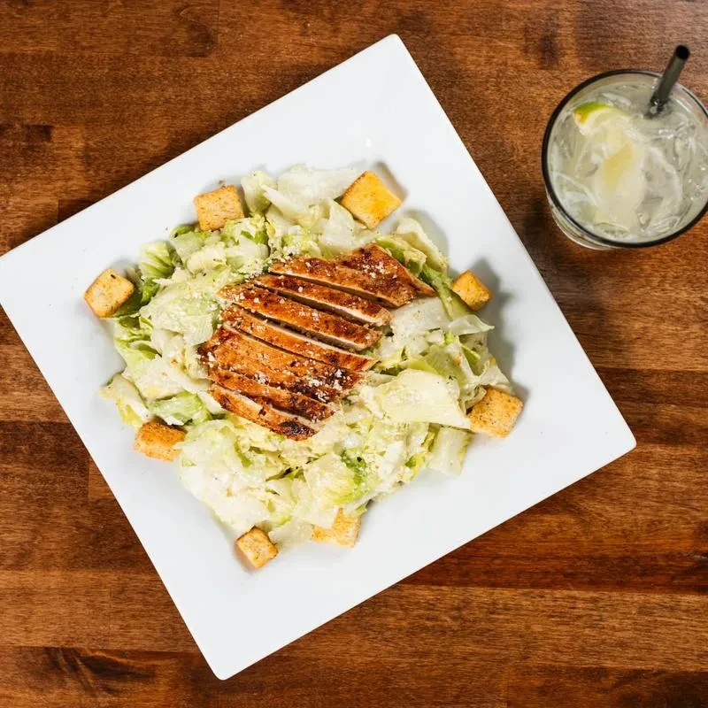 Top-down: Grilled chicken Caesar salad and sparkling drink on wooden table.