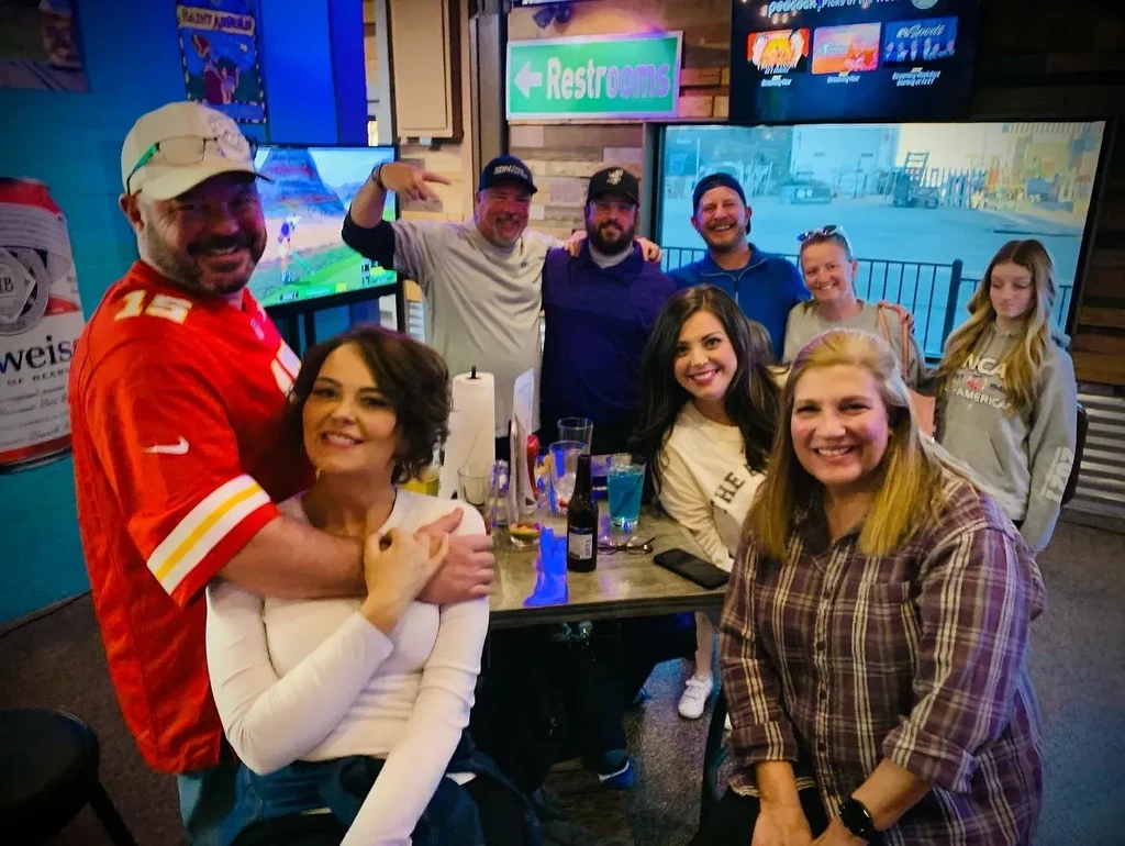 Nine smiling people in a restaurant/sports bar, man in Chiefs jersey.