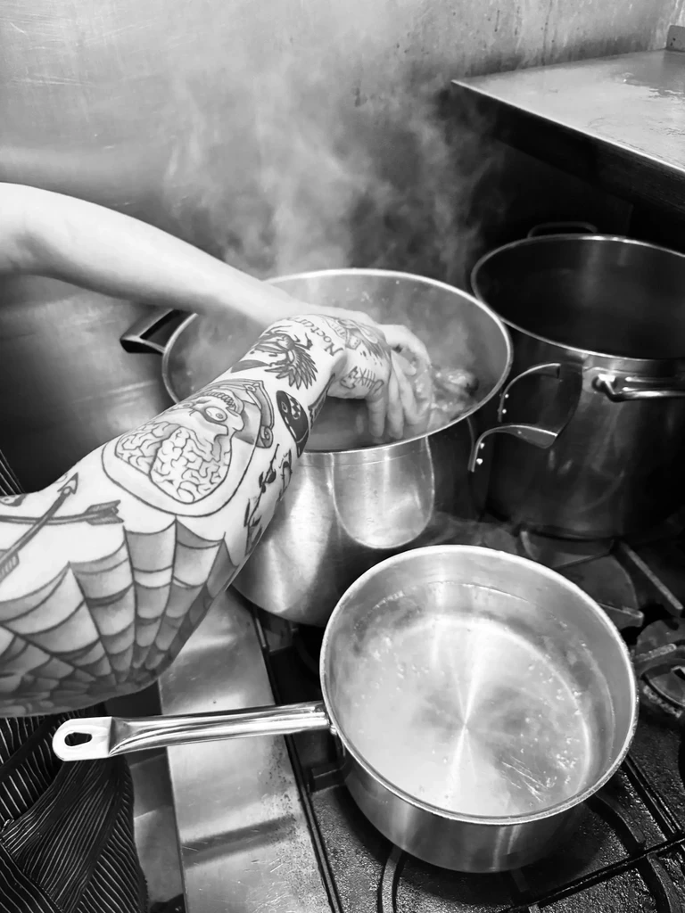 Tattooed arm stirring a steaming pot in a professional kitchen.