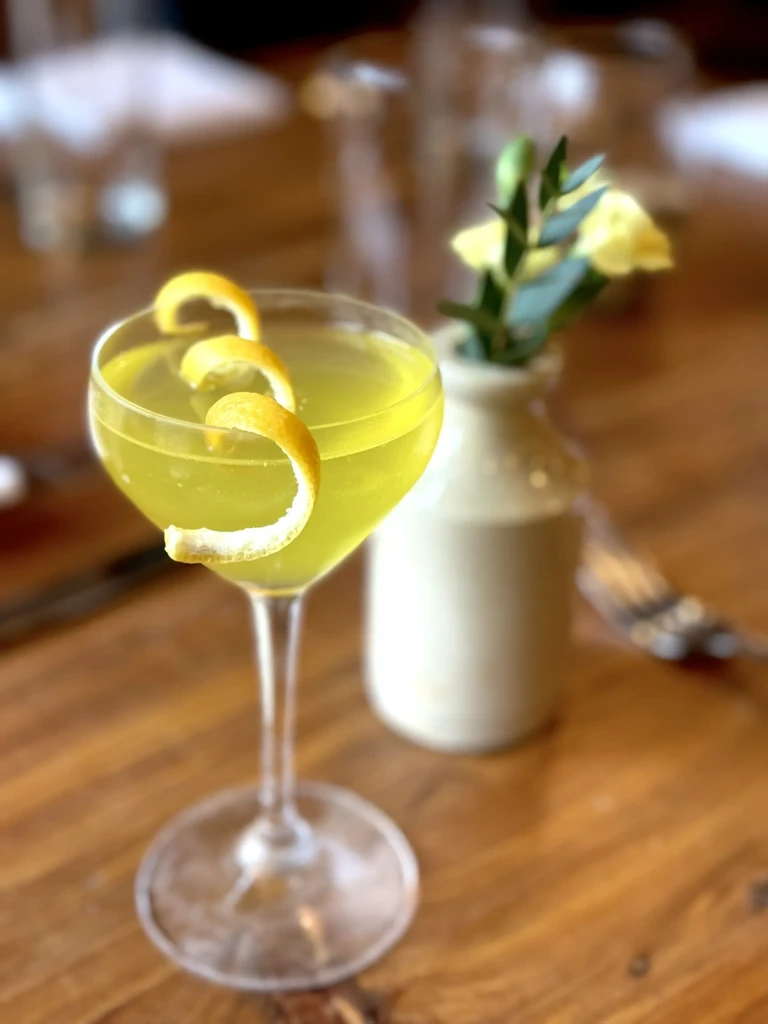 Yellow-green cocktail in coupe glass with lemon peels on a wooden table.