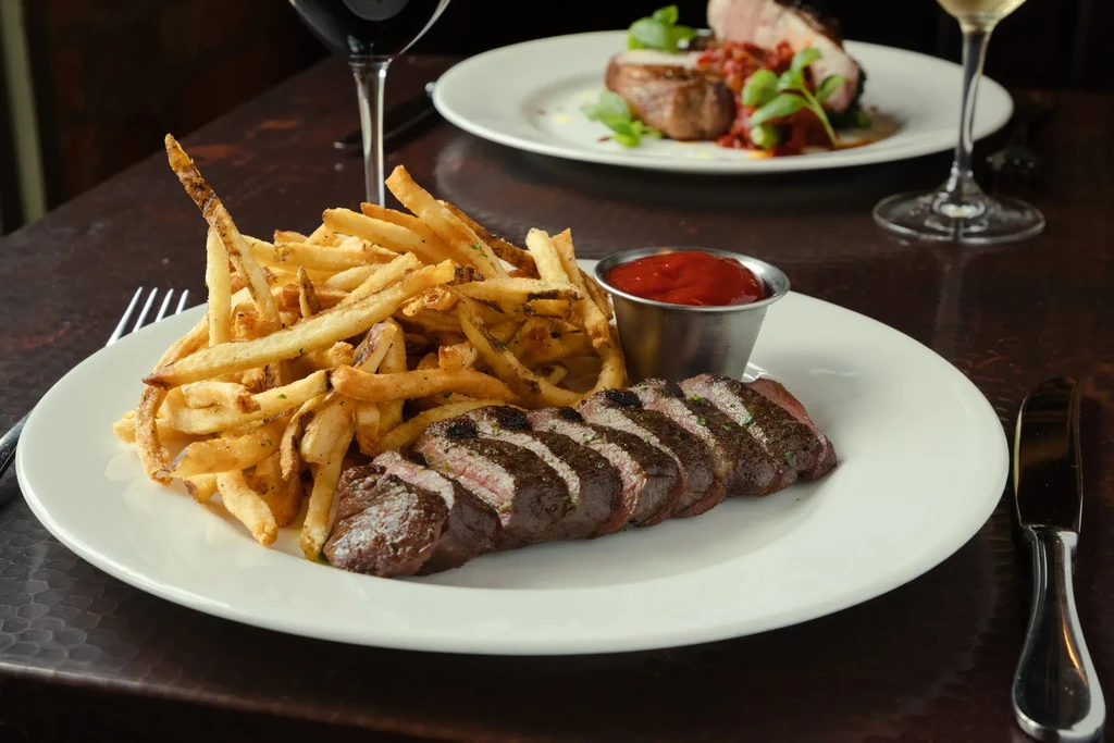 Two plates of steak, fries, and wine on a dark restaurant table.