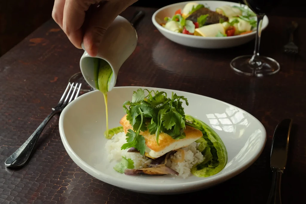 Hand pouring green sauce over fish, rice, and cilantro on a plate.