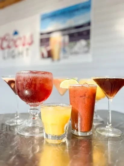 Six colorful drinks on a reflective bar with blurred brewery background.