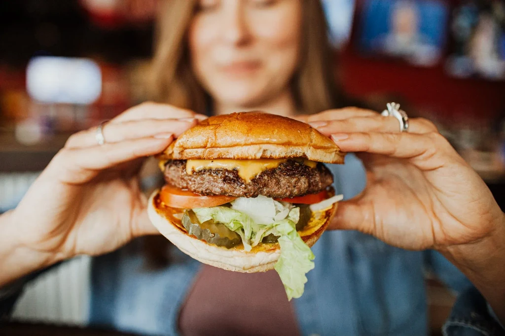 Hands holding a hamburger with beef patty, cheese, and vegetables.