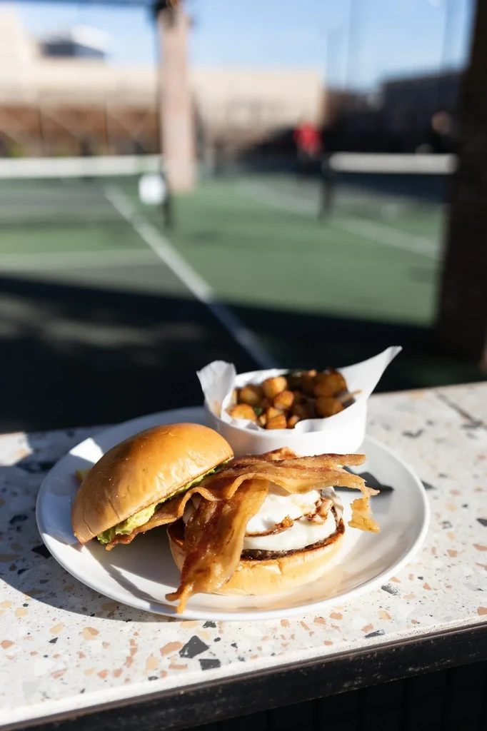 Egg and bacon sandwich, potatoes on white plate, blurred court background.