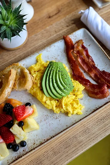 Close-up of a breakfast platter with scrambled eggs, bacon, toast, fruit.