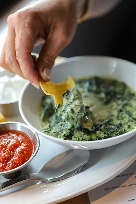 Hand dipping golden chip into creamy green spinach dip.