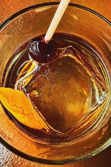 Dark amber cocktail with cherry, orange peel, and square ice cube.