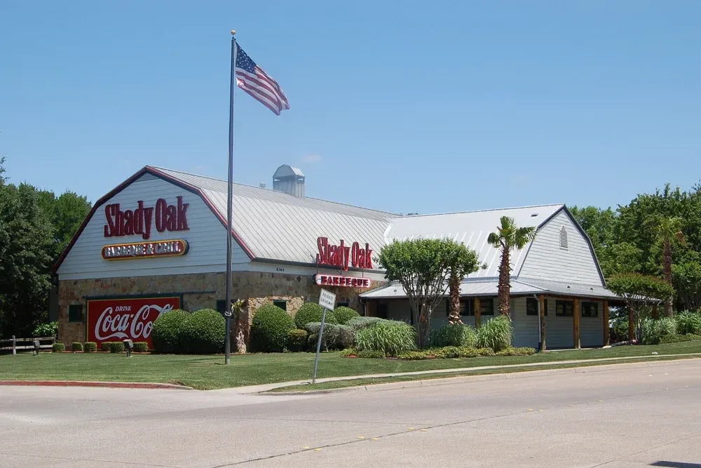 Barn-style Shady Oak Barbecue & Grill with flag, palm trees, blue sky.